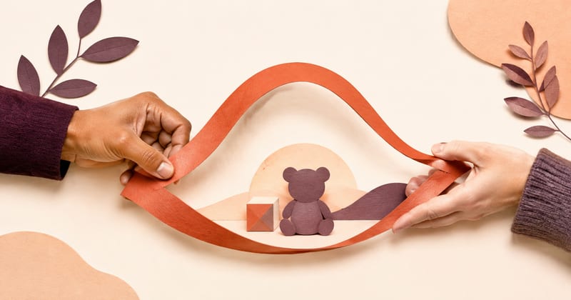Two hands drawing a paper ribbon into a gentle boundary around a teddy bear and a stacking block
