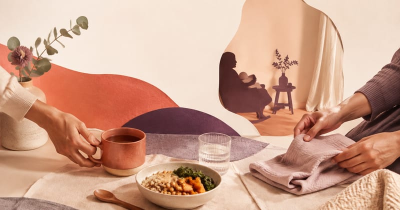 One person passing a mug of tea across a table while another folds a muslin cloth, with a parent feeding a baby in the window behind
