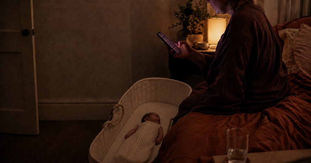 A dimly lit bedroom before dawn. A parent sits on the edge of the bed with a glowing phone in one hand, while their baby sleeps on their back in a bassinet beside them.